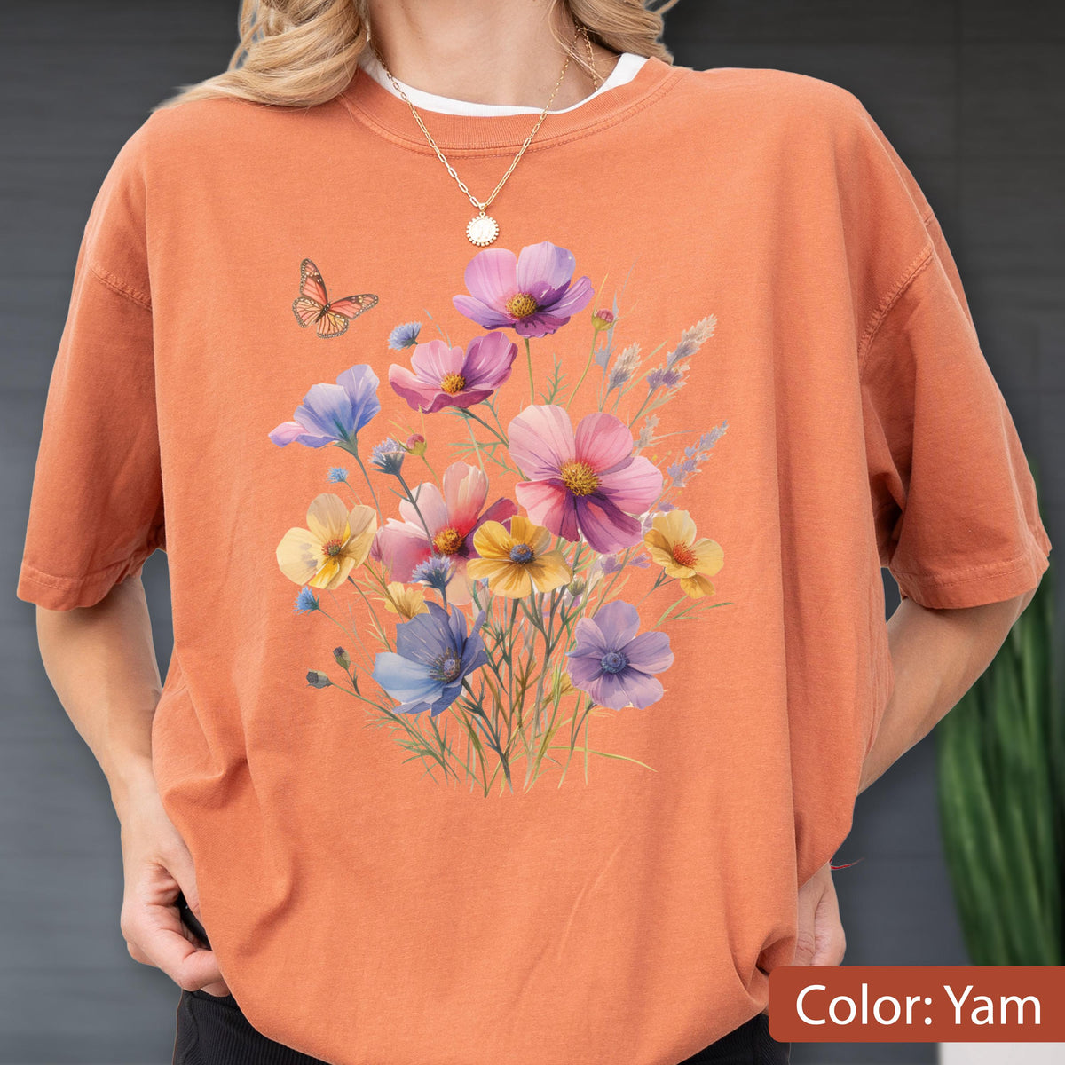 May include: A short-sleeved crew neck t-shirt in a solid burnt orange color. The front features a printed graphic of a colorful wildflower bouquet in shades of pink, purple, yellow, and blue, accompanied by a small orange butterfly.