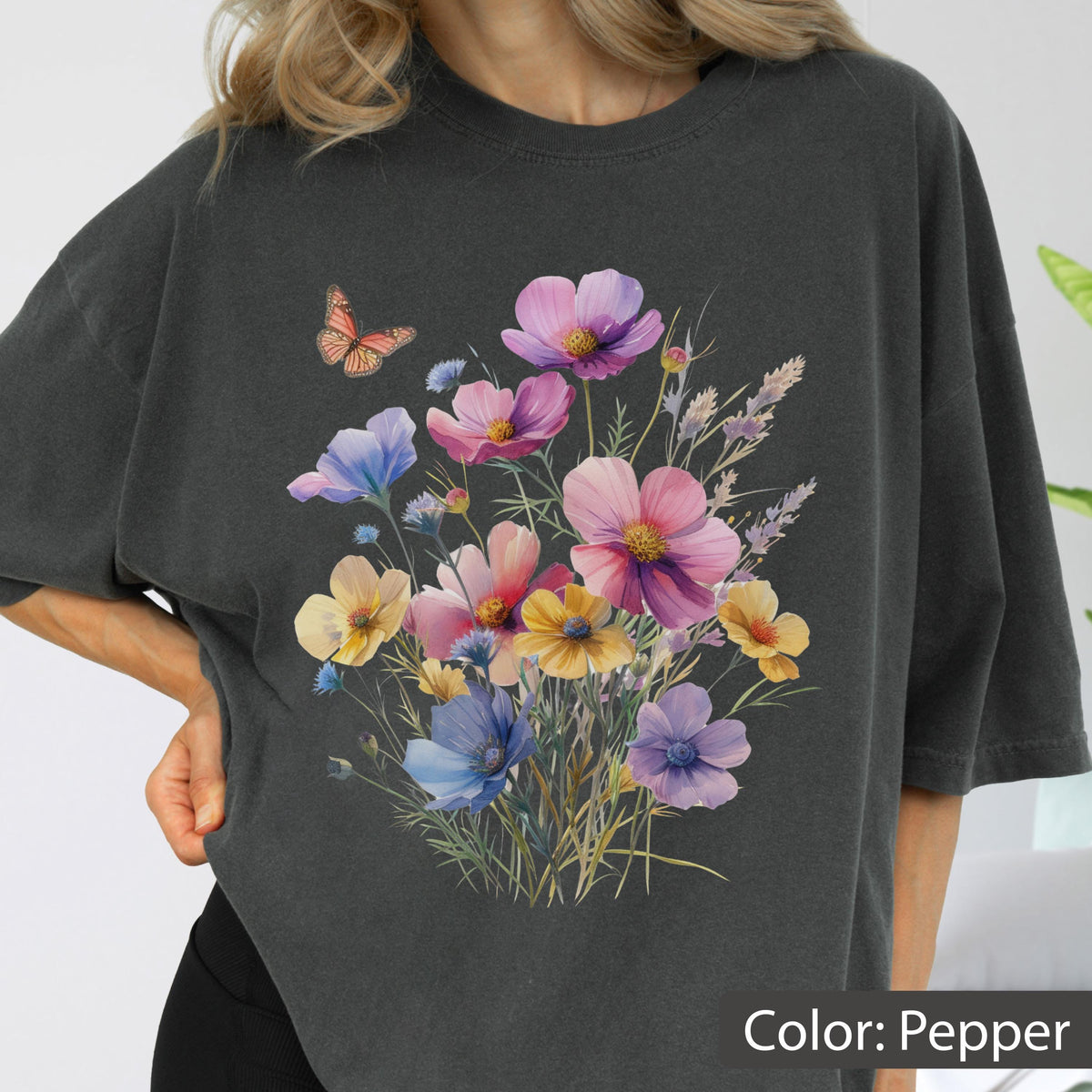 May include: A charcoal gray t-shirt featuring a central graphic of a colorful wildflower bouquet with pink, purple, blue, and yellow petals, accompanied by a small orange butterfly. The shirt has a crew neckline and short sleeves in a relaxed fit.