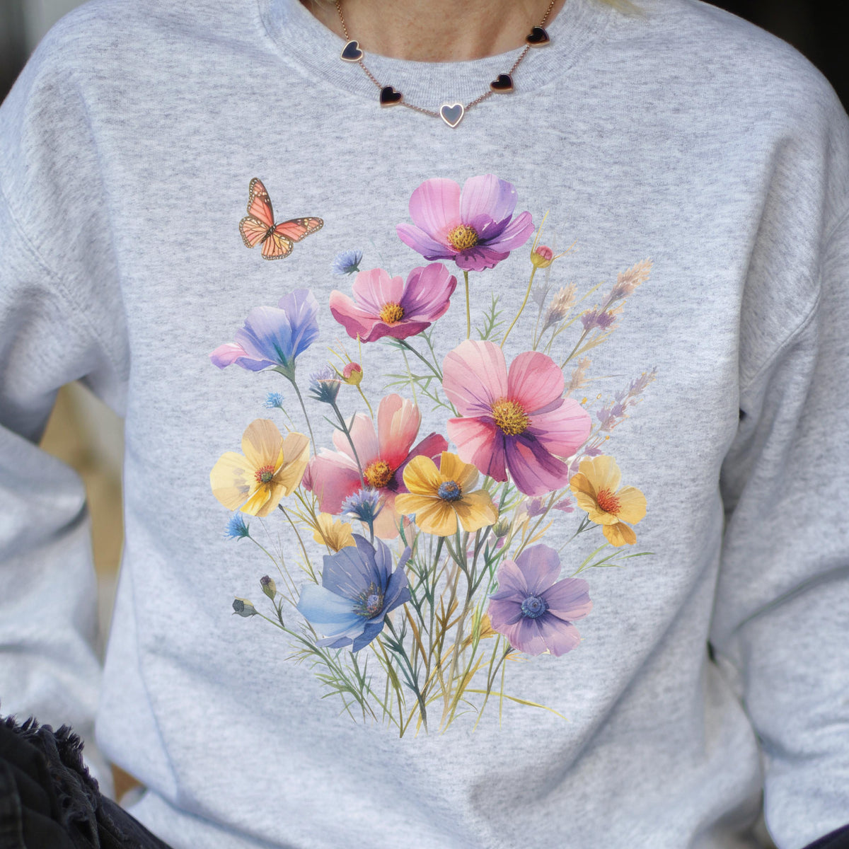 May include: A light gray sweatshirt featuring a watercolor floral design with pink, yellow, and blue flowers. A small orange butterfly is on the left side of the sweatshirt. The floral design is centered on the front.