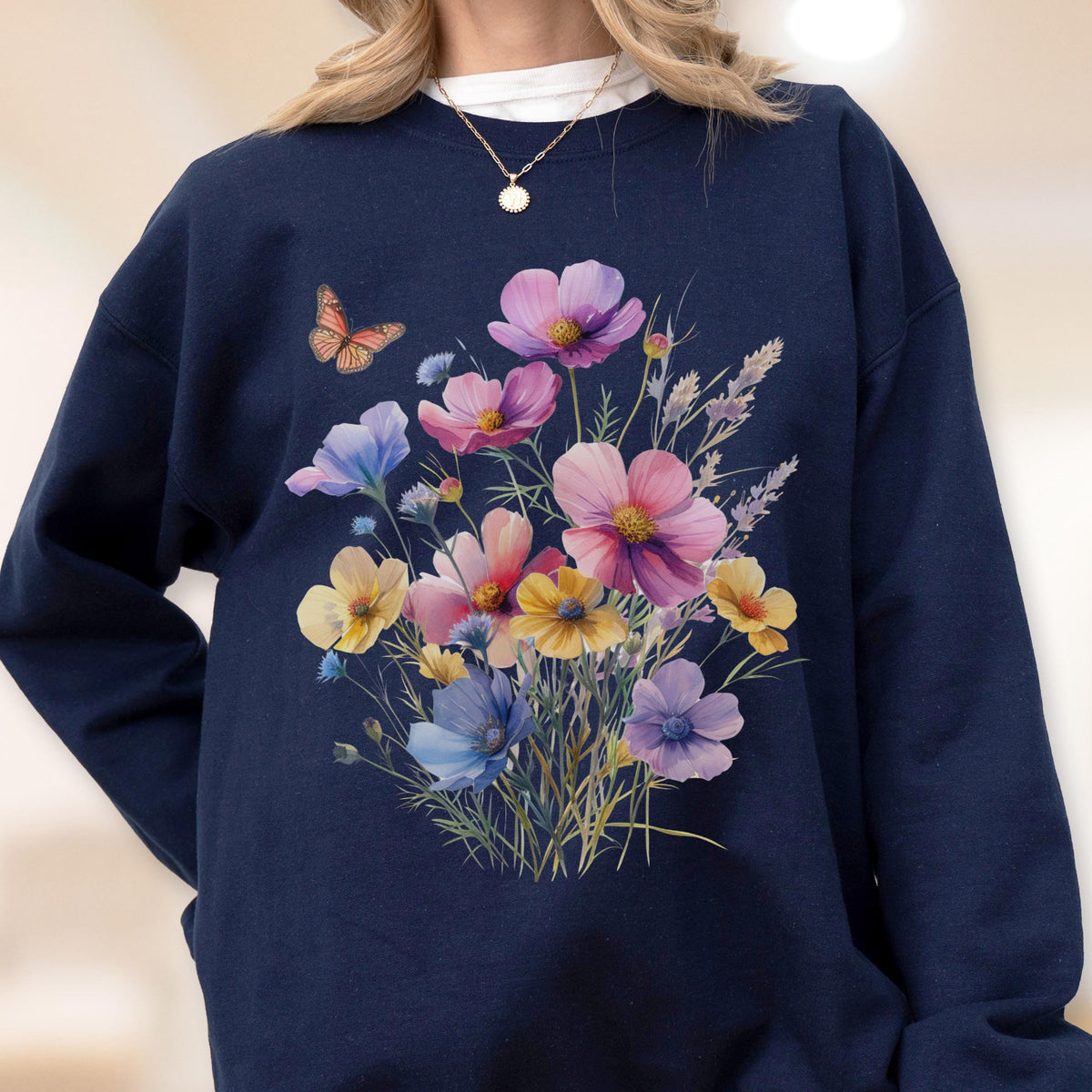 May include: Navy blue sweatshirt featuring a watercolor floral design. The design includes a bouquet of pink, purple, yellow, and blue flowers, with a small butterfly. The sweatshirt has a crew neck and long sleeves.