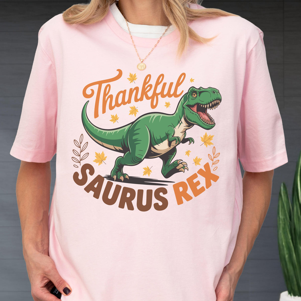 May include: A light pink t-shirt featuring a green Tyrannosaurus Rex dinosaur with the words "Thankful Saurus Rex" in orange and brown. The design includes fall leaves and decorative elements, perfect for the autumn season.