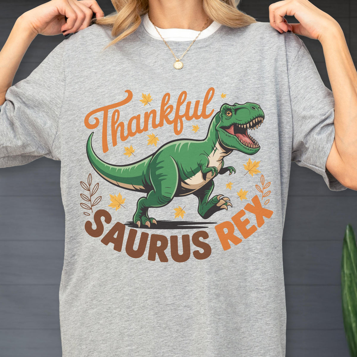 May include: Gray t-shirt with a green dinosaur graphic and the text "Thankful Saurus Rex" in orange and brown. The design includes fall leaves and a decorative leaf sprig. The shirt has a white collar.