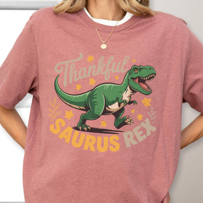 May include: A dusty rose t-shirt featuring a green Tyrannosaurus Rex illustration with the words "Thankful Saurus Rex" in a playful font. The design includes fall leaves and stars.