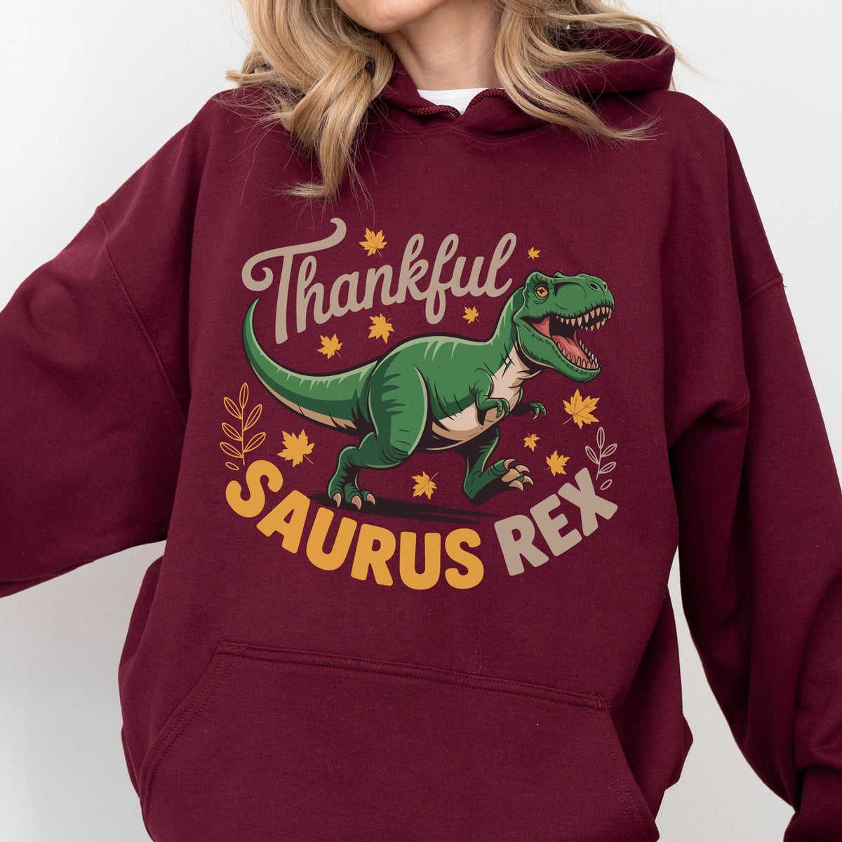 May include: A maroon hoodie featuring a green Tyrannosaurus Rex illustration with the text "Thankful Saurus Rex" in a playful font. The design includes fall leaves and decorative elements, perfect for the autumn season.