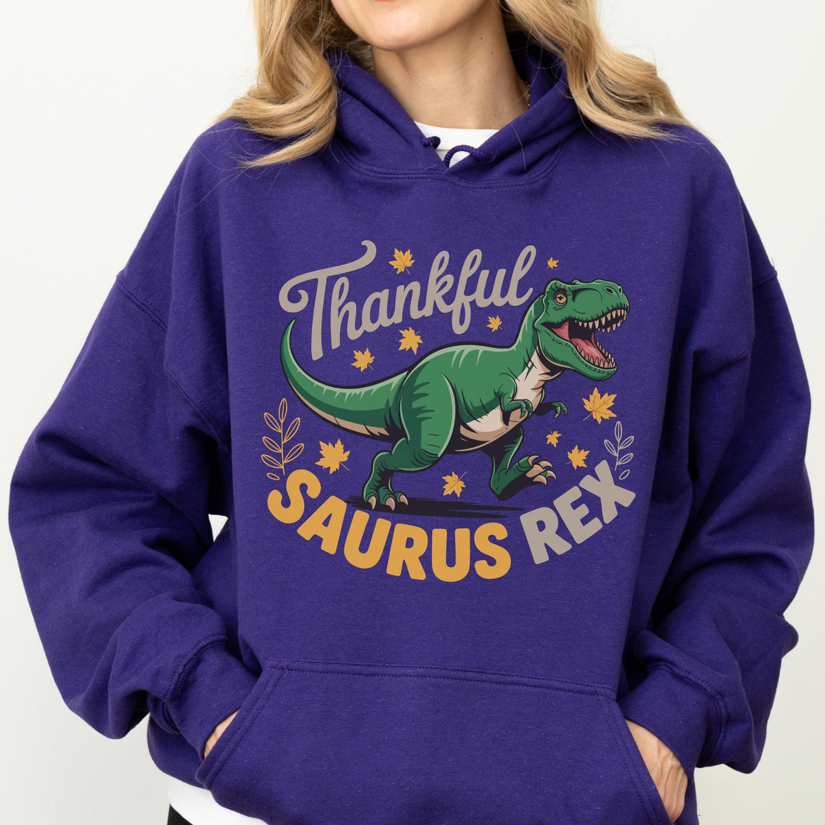 May include: A purple hoodie with a graphic of a green Tyrannosaurus Rex and the words "Thankful Saurus Rex" in a playful font. The design includes fall leaves. The hoodie has a drawstring hood and a front pocket.
