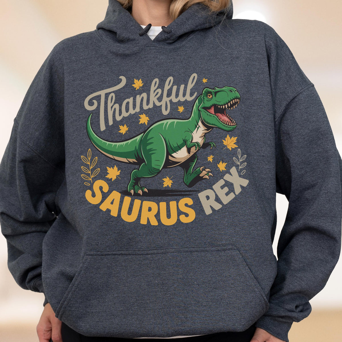 May include: Dark gray hoodie featuring a green Tyrannosaurus Rex illustration with the text "Thankful Saurus Rex". The design includes fall leaves and a decorative border, perfect for the autumn season.