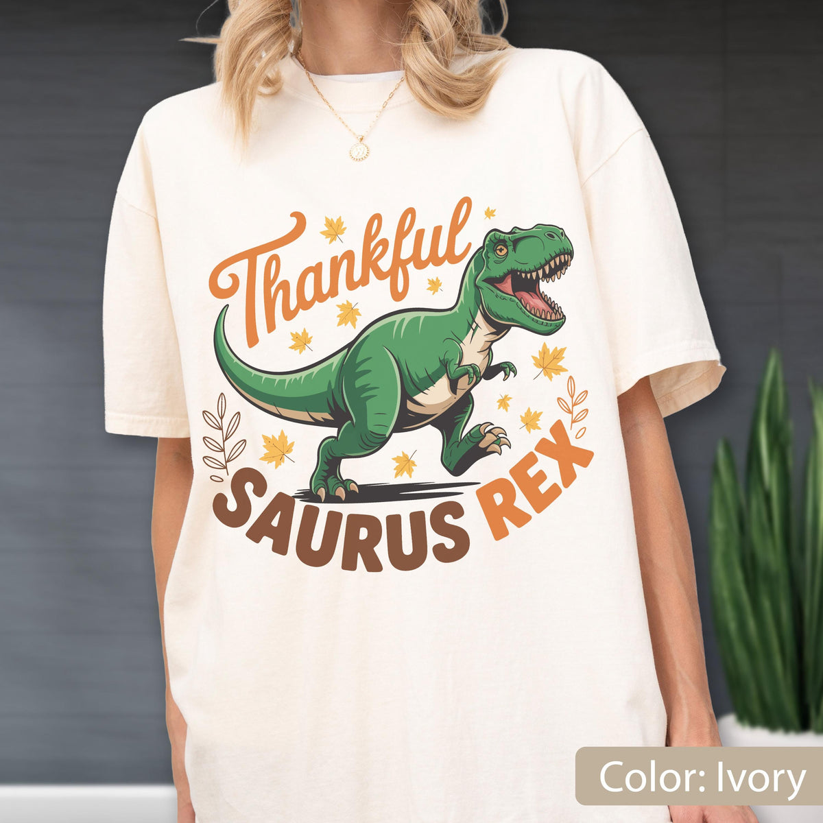 May include: Ivory-colored t-shirt featuring a green Tyrannosaurus Rex illustration with the text "Thankful Saurus Rex" in orange and brown. The design includes fall leaves and a leaf sprig.