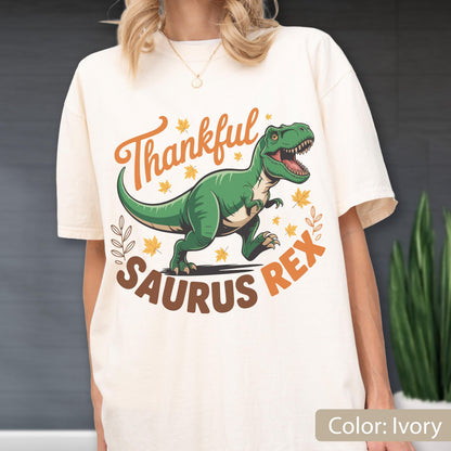 May include: Ivory-colored t-shirt featuring a green Tyrannosaurus Rex illustration with the text "Thankful Saurus Rex" in orange and brown. The design includes fall leaves and a leaf sprig.