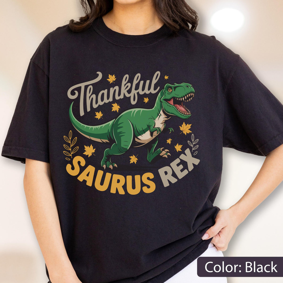 May include: Black t-shirt with a green dinosaur graphic and the text "Thankful Saurus Rex". The design includes fall leaves and a decorative border. The shirt is a comfortable, casual style.