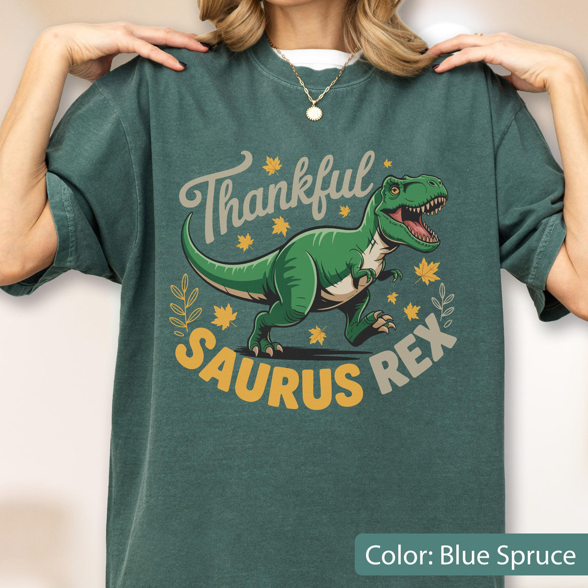 May include: Blue spruce t-shirt with a graphic of a green Tyrannosaurus Rex and the words "Thankful Saurus Rex". The design includes fall leaves and a decorative wreath. The shirt is a comfortable, casual style.