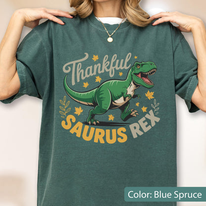 May include: Blue spruce t-shirt with a graphic of a green Tyrannosaurus Rex and the words "Thankful Saurus Rex". The design includes fall leaves and a decorative wreath. The shirt is a comfortable, casual style.