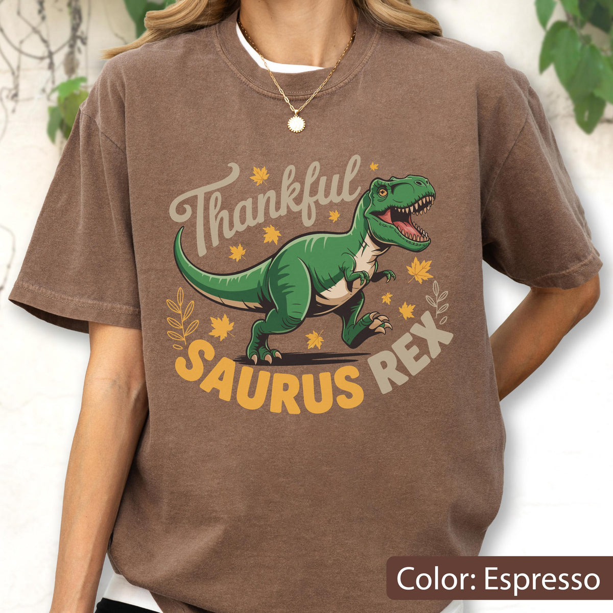 May include: Espresso-colored t-shirt featuring a green Tyrannosaurus Rex illustration with the text "Thankful Saurus Rex". The design includes fall leaves and a decorative border, perfect for dinosaur and autumn enthusiasts.