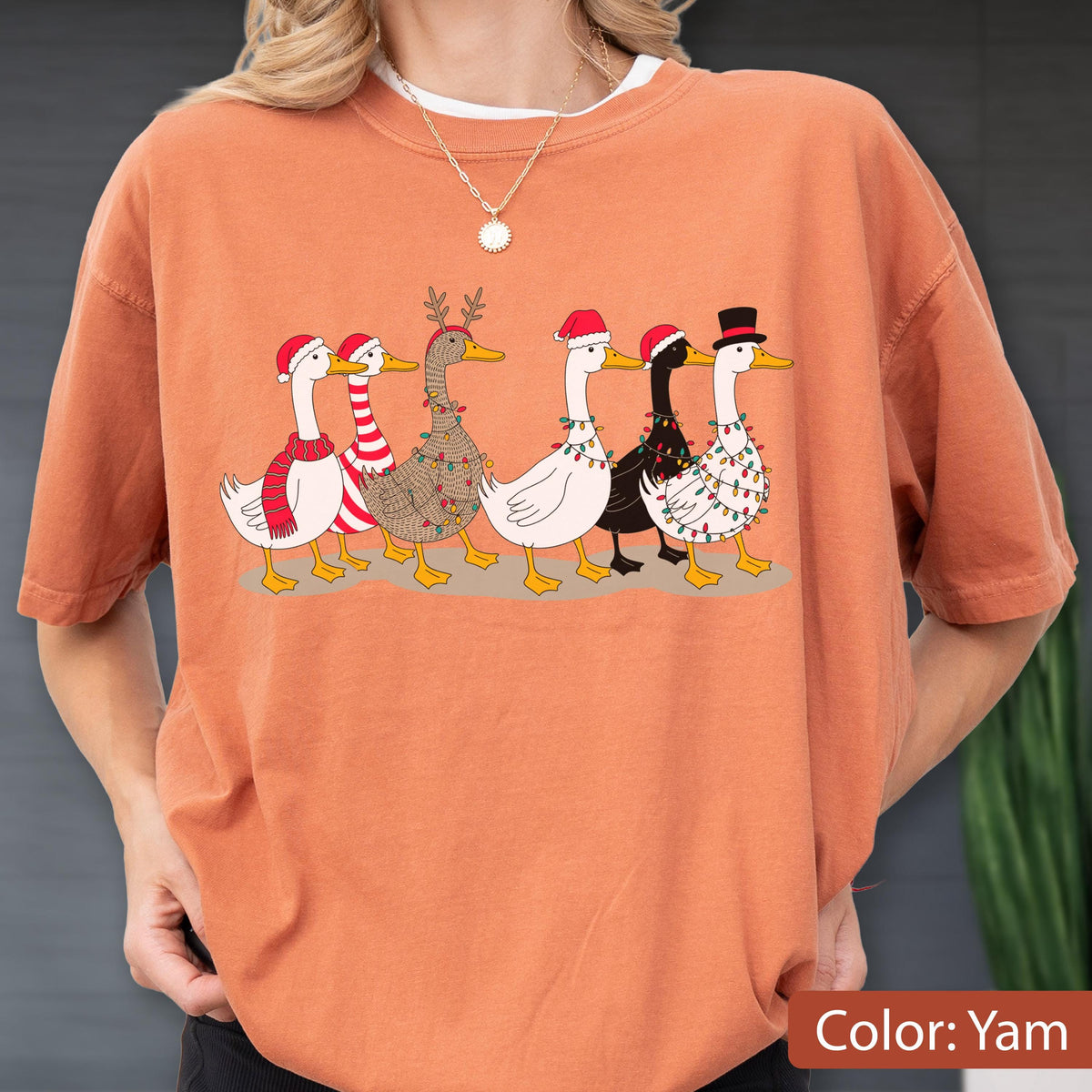 May include: A yam-colored t-shirt featuring a graphic of six ducks in a row. The ducks are decorated for the holidays with Santa hats, a striped scarf, reindeer antlers, and colorful string lights. One duck wears a black top hat.