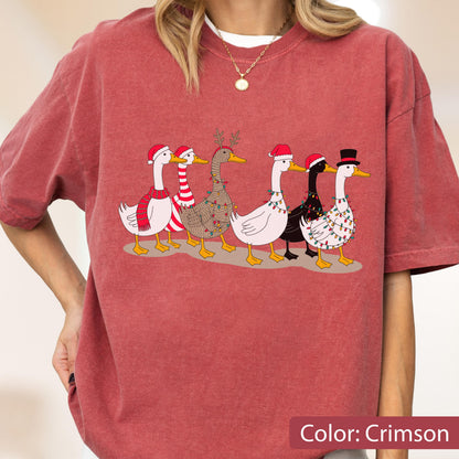May include: A crimson short-sleeve t-shirt featuring a graphic of six geese decorated for the holidays. The geese wear various accessories including Santa hats, a red and white striped scarf, reindeer antlers, and strings of colorful Christmas lights.