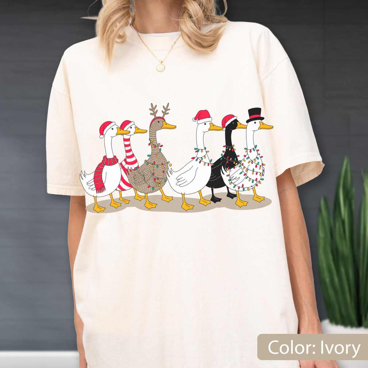 May include: An ivory t-shirt featuring a graphic print of six ducks in a row. The ducks are decorated with Christmas elements including Santa hats, a red and white striped scarf, reindeer antlers, and colorful string lights wrapped around them.
