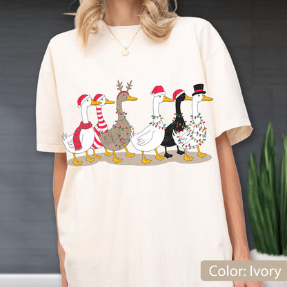 May include: An ivory t-shirt featuring a graphic print of six ducks in a row. The ducks are decorated with Christmas elements including Santa hats, a red and white striped scarf, reindeer antlers, and colorful string lights wrapped around them.