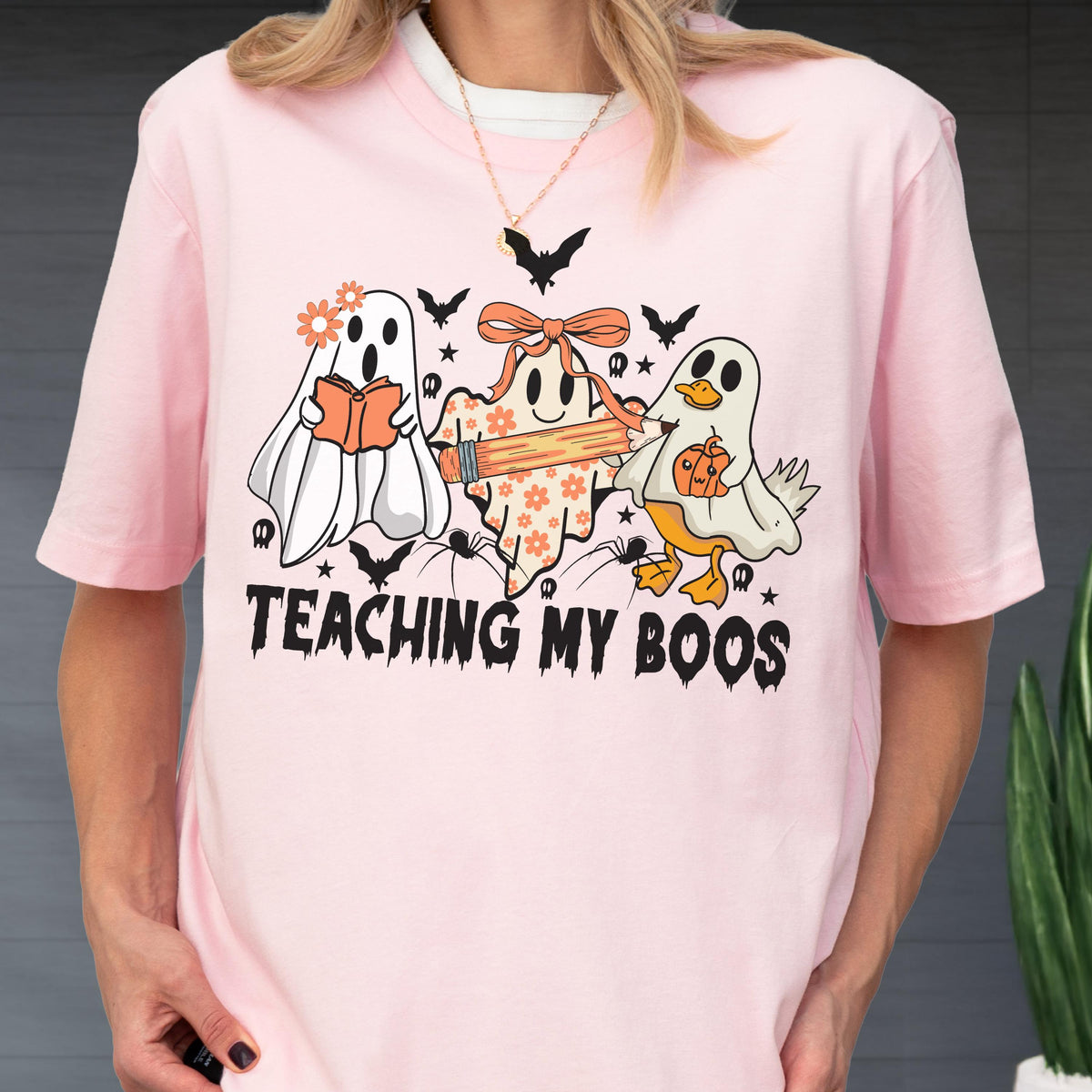 May include: Pink t-shirt with a Halloween-themed graphic. The design features three cartoon ghosts, bats, and the text "TEACHING MY BOOS". One ghost reads a book, another holds a pencil, and the third holds a pumpkin.