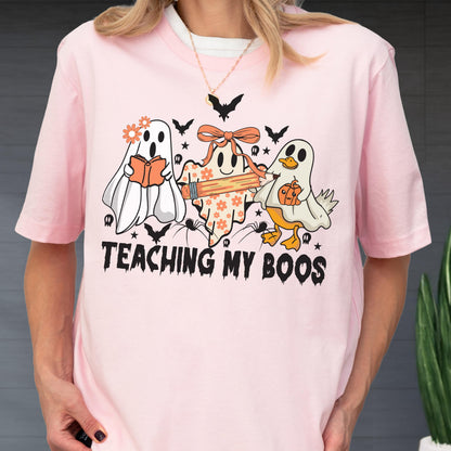 May include: Pink t-shirt with a Halloween-themed graphic. The design features three cartoon ghosts, bats, and the text "TEACHING MY BOOS". One ghost reads a book, another holds a pencil, and the third holds a pumpkin.