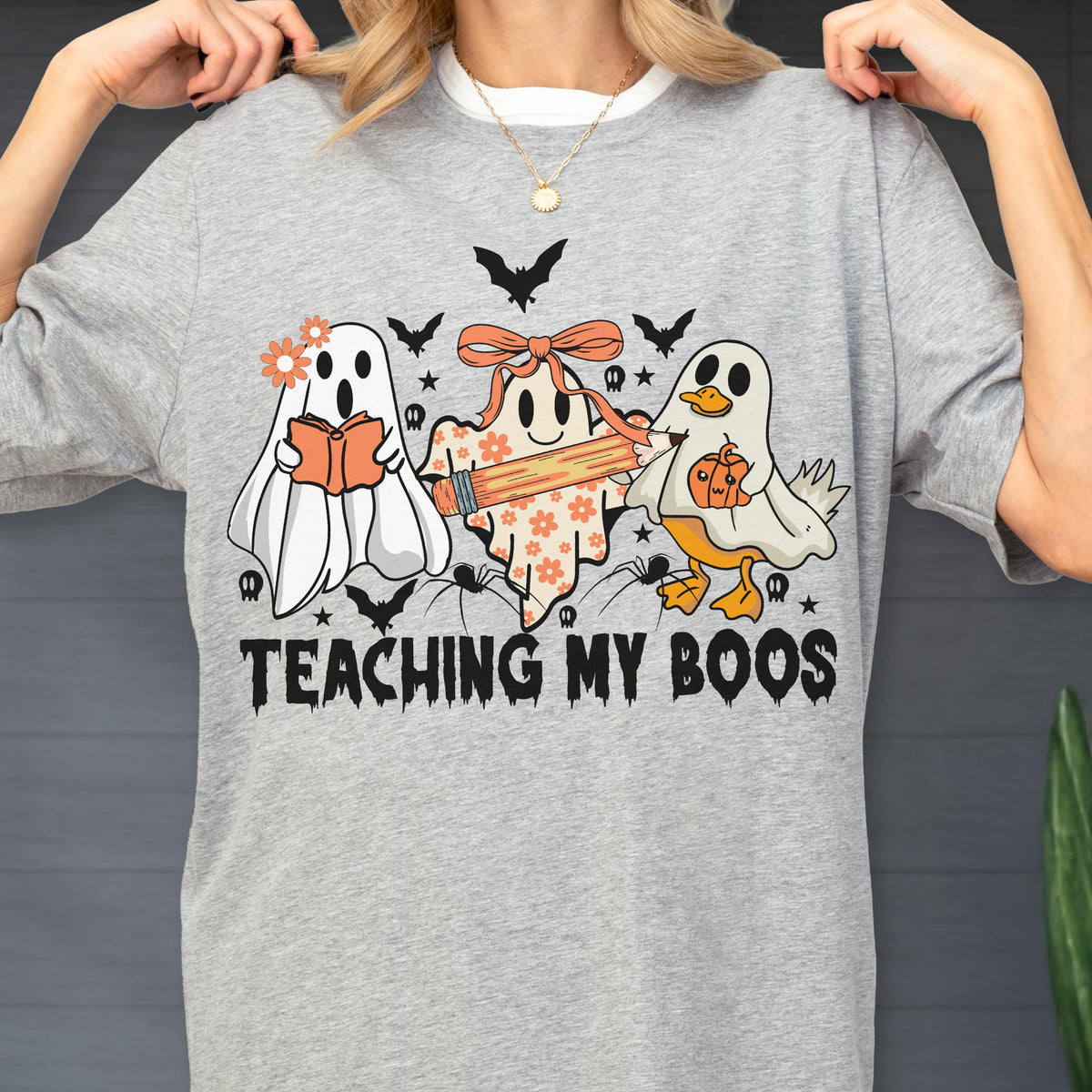 May include: Gray t-shirt featuring a Halloween-themed graphic. The design includes three cartoon ghosts, bats, spiders, and the text "TEACHING MY BOOS". One ghost holds a book, another a pencil, and the third a pumpkin.