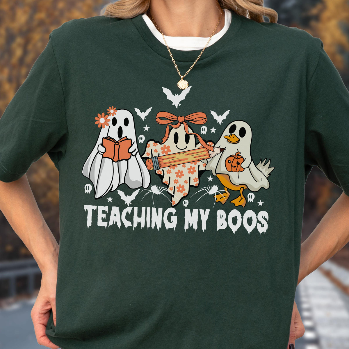 May include: Dark green t-shirt featuring a Halloween-themed graphic. The design includes three cartoon ghosts, one reading a book, one holding a pencil, and one with a pumpkin. The text "TEACHING MY BOOS" is printed below the ghosts.