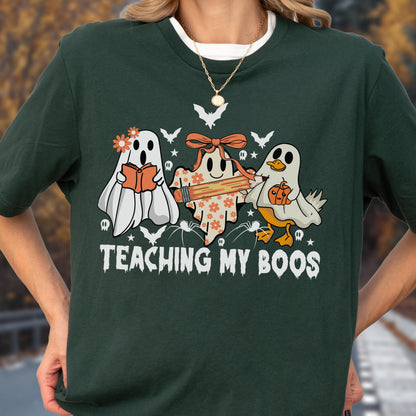 May include: Dark green t-shirt featuring a Halloween-themed graphic. The design includes three cartoon ghosts, one reading a book, one holding a pencil, and one with a pumpkin. The text "TEACHING MY BOOS" is printed below the ghosts.