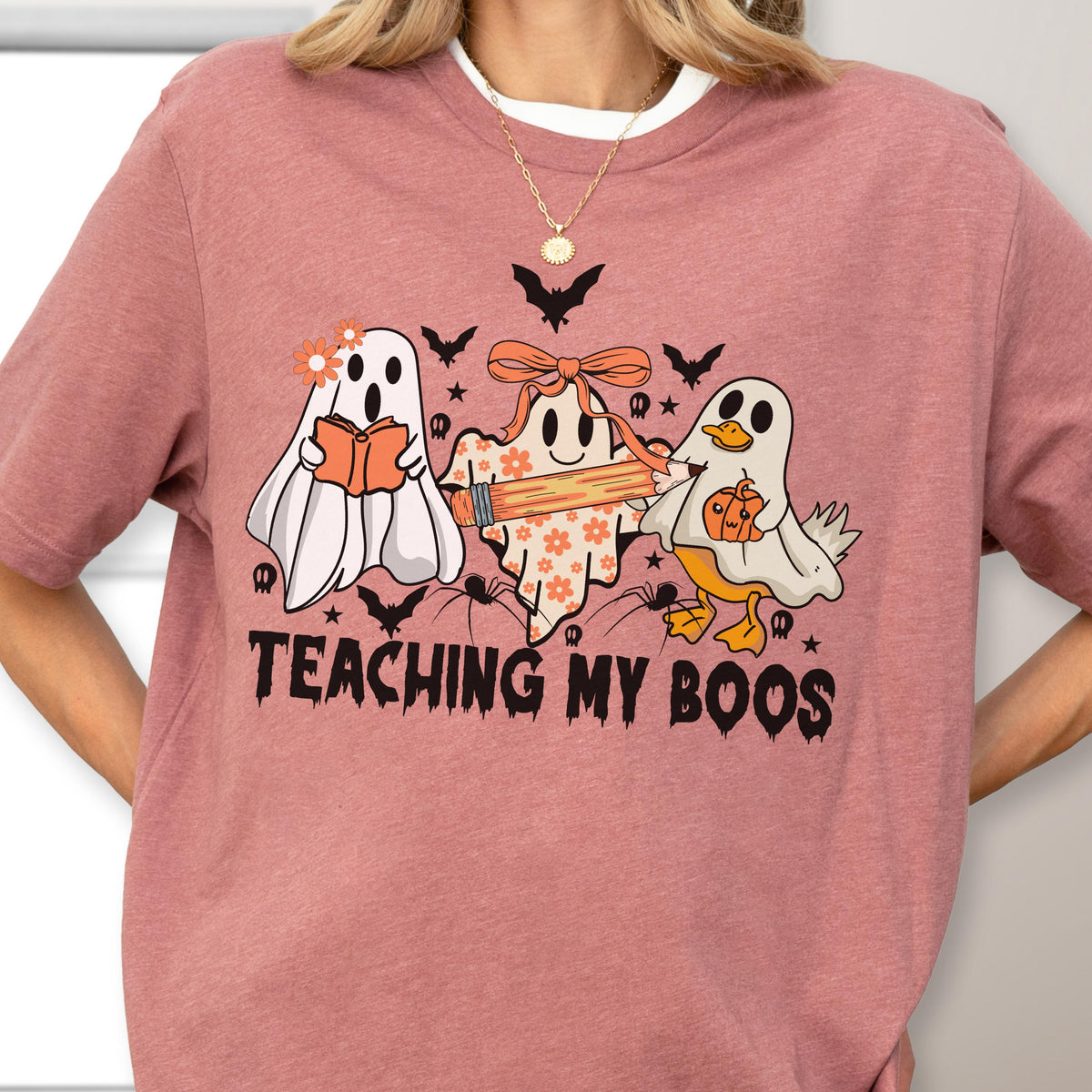 May include: A dusty rose t-shirt featuring a Halloween-themed graphic. The design includes three cartoon ghosts, bats, spiders, and the text "TEACHING MY BOOS" in black. One ghost reads a book, another holds a pencil, and the third has a pumpkin.
