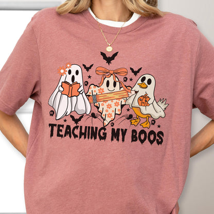 May include: A dusty rose t-shirt featuring a Halloween-themed graphic. The design includes three cartoon ghosts, bats, spiders, and the text "TEACHING MY BOOS" in black. One ghost reads a book, another holds a pencil, and the third has a pumpkin.