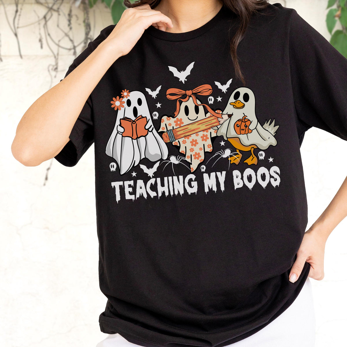 May include: Black t-shirt with a Halloween-themed graphic. The design features three cartoon ghosts, bats, pumpkins, and the text "TEACHING MY BOOS". The ghosts are holding a book, a sign, and a pumpkin.