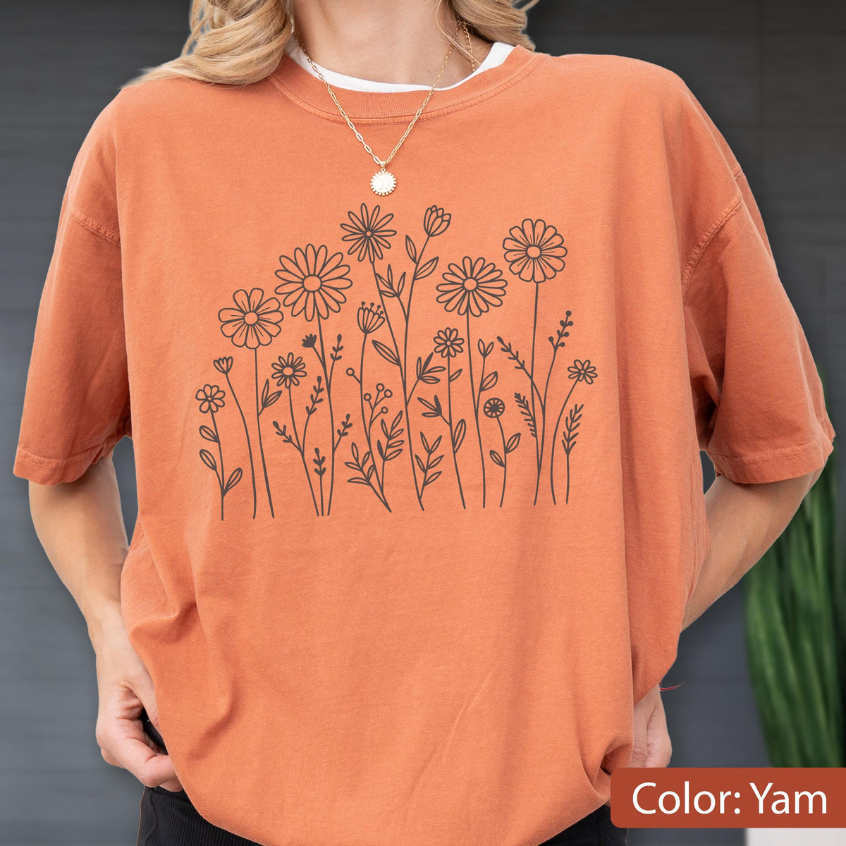 May include: A short-sleeve crew neck t-shirt in a solid burnt orange color. The front of the shirt features a black line art illustration of various wildflowers and daisies arranged in a cluster. A thin gold chain necklace is worn around the neck.