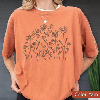 May include: A short-sleeve crew neck t-shirt in a solid burnt orange color. The front of the shirt features a black line art illustration of various wildflowers and daisies arranged in a cluster. A thin gold chain necklace is worn around the neck.