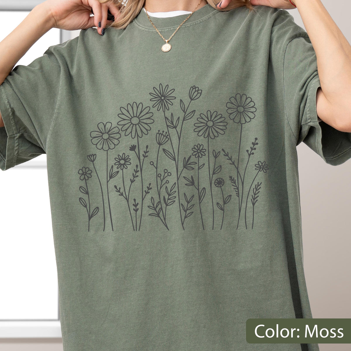May include: A moss green short-sleeve t-shirt featuring a dark gray line art illustration of various wildflowers and stems across the front. The shirt has a crew neckline and a relaxed fit, showcasing a botanical design with daisies and foliage.