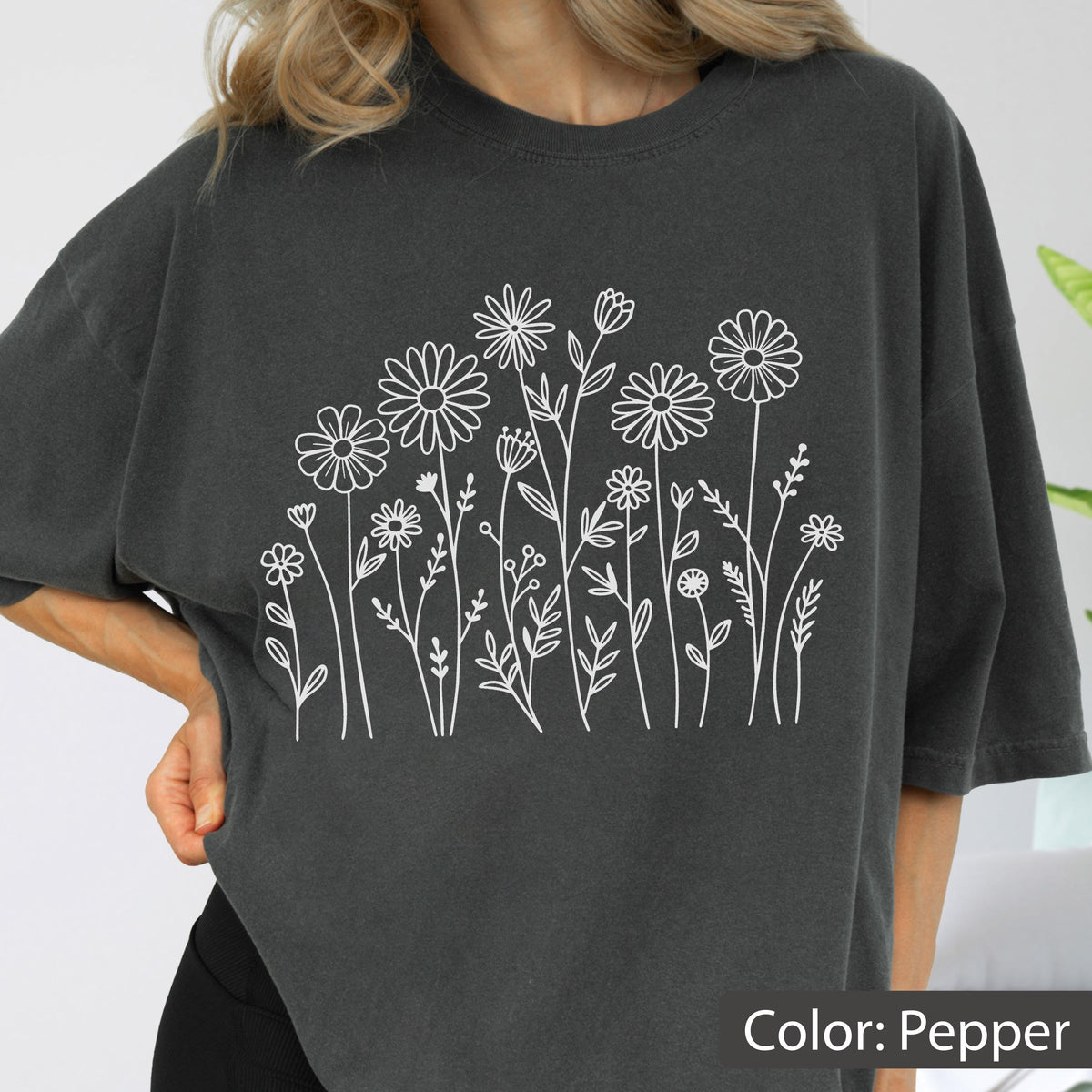 May include: A charcoal gray short-sleeved t-shirt featuring a white line art graphic of various wildflowers and stems across the center. The shirt has a crew neckline and a relaxed fit, with the color labeled as pepper in the bottom corner.