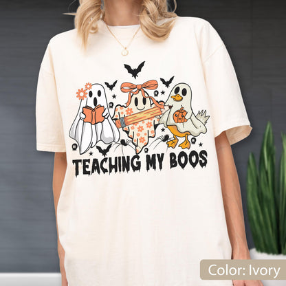 May include: Ivory-colored t-shirt featuring a Halloween-themed graphic. The design includes three cartoon ghosts, bats, and the phrase "TEACHING MY BOOS." One ghost reads a book, another holds a pencil, and the third carries a pumpkin.