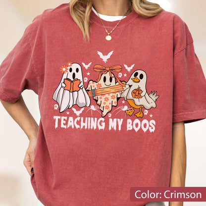 May include: Crimson-colored t-shirt featuring a graphic of three cartoon ghosts. One ghost reads a book, another holds a pencil, and the third has a pumpkin. The text "TEACHING MY BOOS" is below the ghosts. Bats and stars are also in the design.