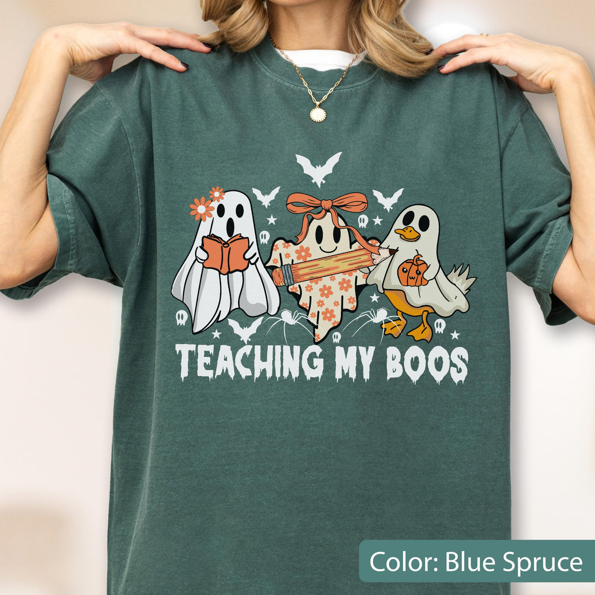 May include: Blue spruce t-shirt with a Halloween-themed graphic. The design features three cartoon ghosts, one reading a book, one holding a pencil, and one with a pumpkin. The text "TEACHING MY BOOS" is printed below the ghosts.