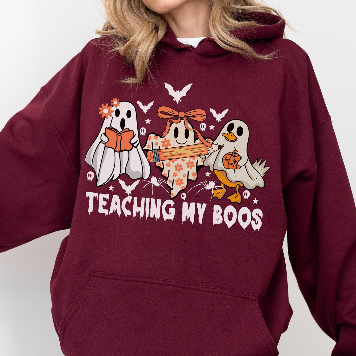 May include: A maroon hooded sweatshirt featuring a graphic of three ghosts, one holding a book, one with a pencil, and one with a pumpkin. The text "TEACHING MY BOOS" is printed below the ghosts, surrounded by small bats, stars, and spiders.