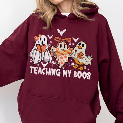 May include: A maroon hooded sweatshirt featuring a graphic of three ghosts, one holding a book, one with a pencil, and one with a pumpkin. The text "TEACHING MY BOOS" is printed below the ghosts, surrounded by small bats, stars, and spiders.