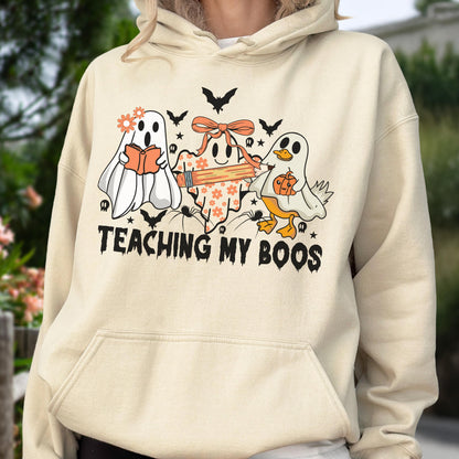 May include: A cream-colored hooded sweatshirt featuring a graphic print of three ghosts, one wearing a flower crown and reading a book, one with a bow holding a pencil, and one dressed as a duck holding a pumpkin, with the text TEACHING MY BOOS.