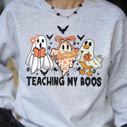 May include: A light gray crewneck sweatshirt featuring a graphic of three ghosts, one holding an orange book, one holding a pencil, and one holding a pumpkin. The text "TEACHING MY BOOS" is printed in black below the ghosts, surrounded by small bats.