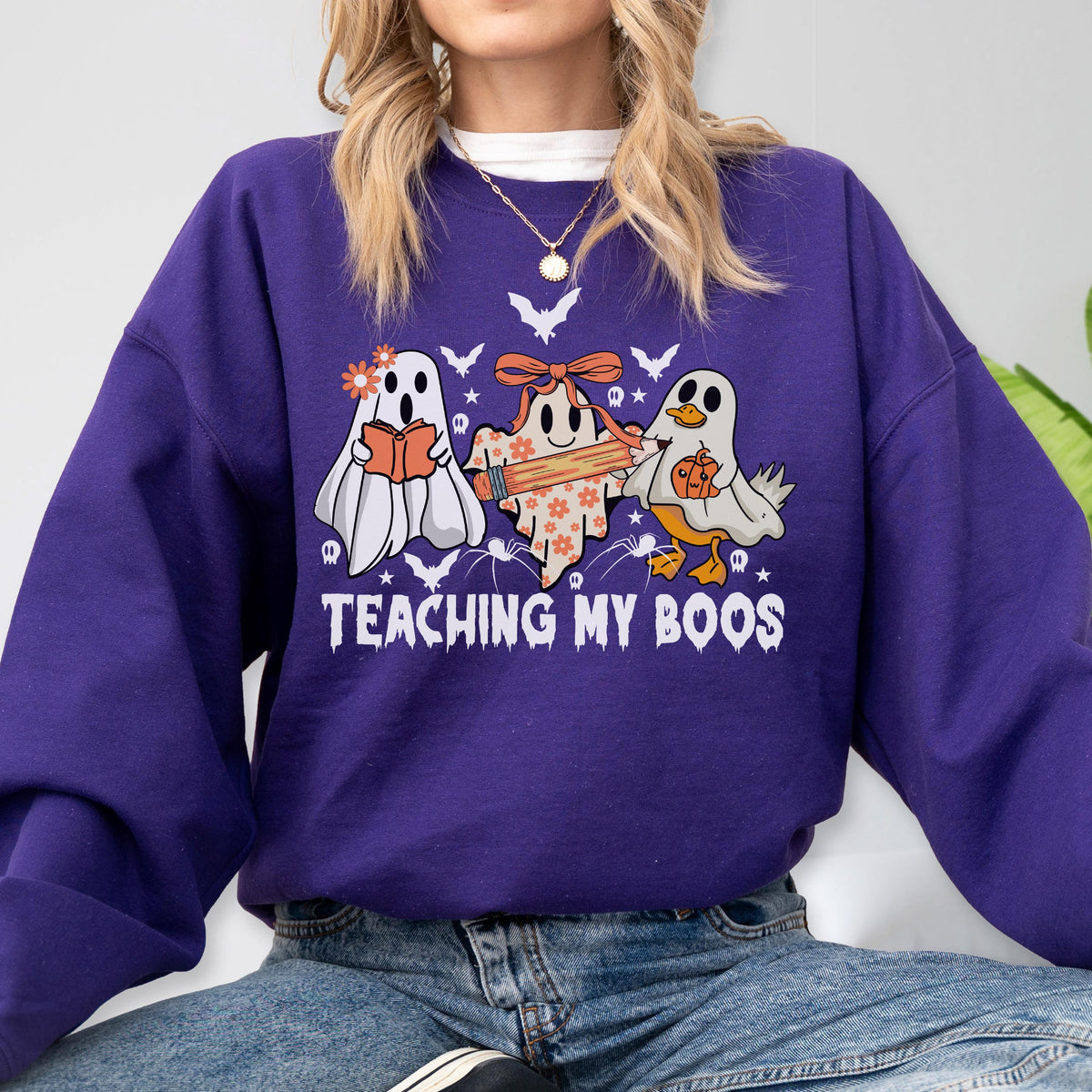 May include: A purple long-sleeve sweatshirt featuring a graphic of three ghosts, one holding a book, one holding a pencil, and one holding a pumpkin. The text "TEACHING MY BOOS" is printed in white below the ghosts, surrounded by small bats and stars.