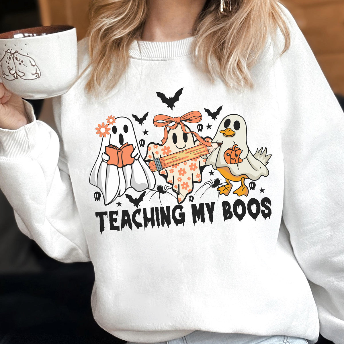 May include: A white crewneck sweatshirt featuring a graphic of three ghosts, one holding an orange book, one with a bow holding a pencil, and one duck ghost holding a pumpkin. The text "TEACHING MY BOOS" is printed in black below the ghosts.