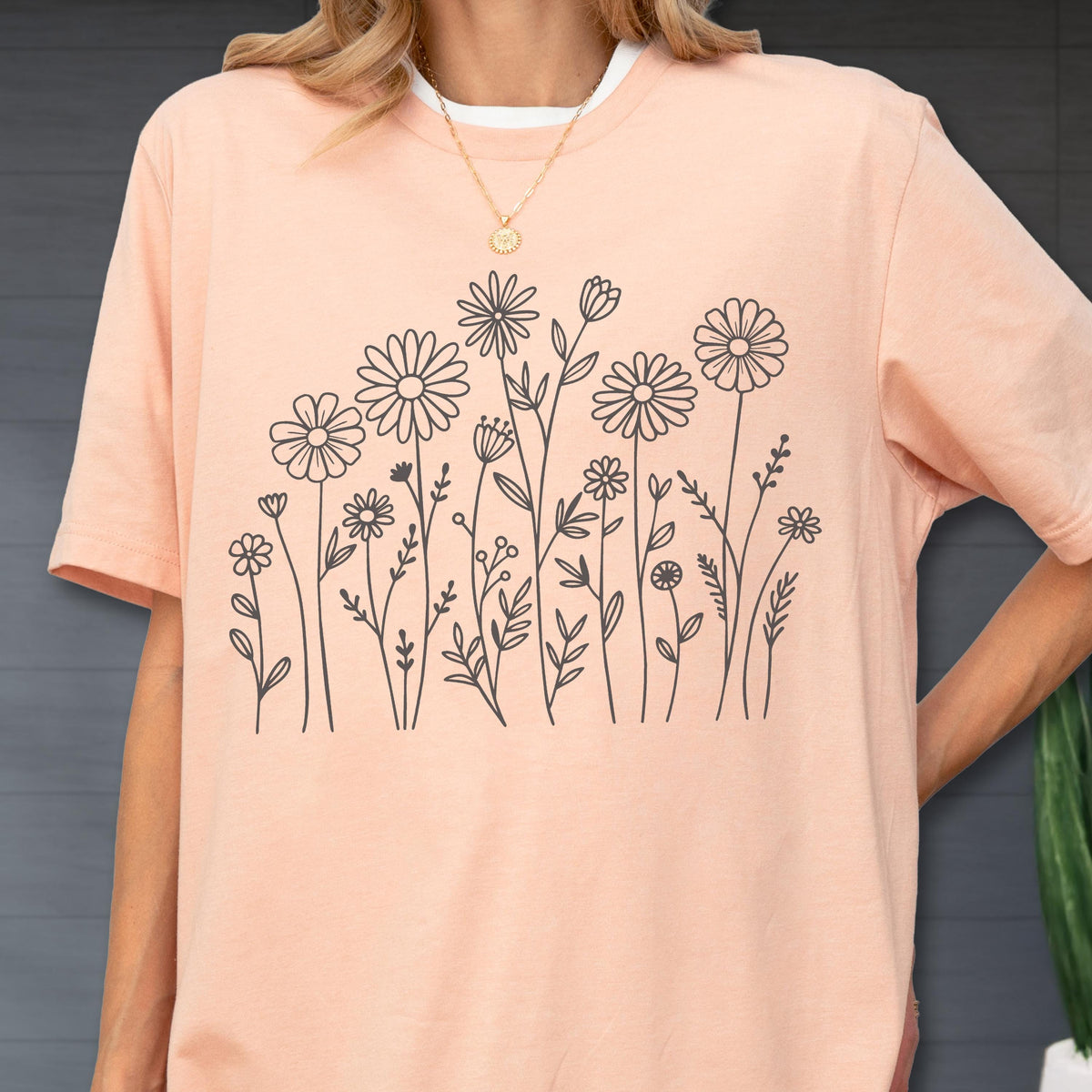 May include: A peach-colored short-sleeve t-shirt featuring a black line art graphic of various wildflowers and stems across the front. The shirt has a crew neckline and a relaxed fit, showcasing a simple botanical illustration of daisies and foliage.