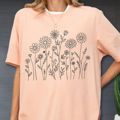 May include: A peach-colored short-sleeve t-shirt featuring a black line art graphic of various wildflowers and stems across the front. The shirt has a crew neckline and a relaxed fit, showcasing a simple botanical illustration of daisies and foliage.