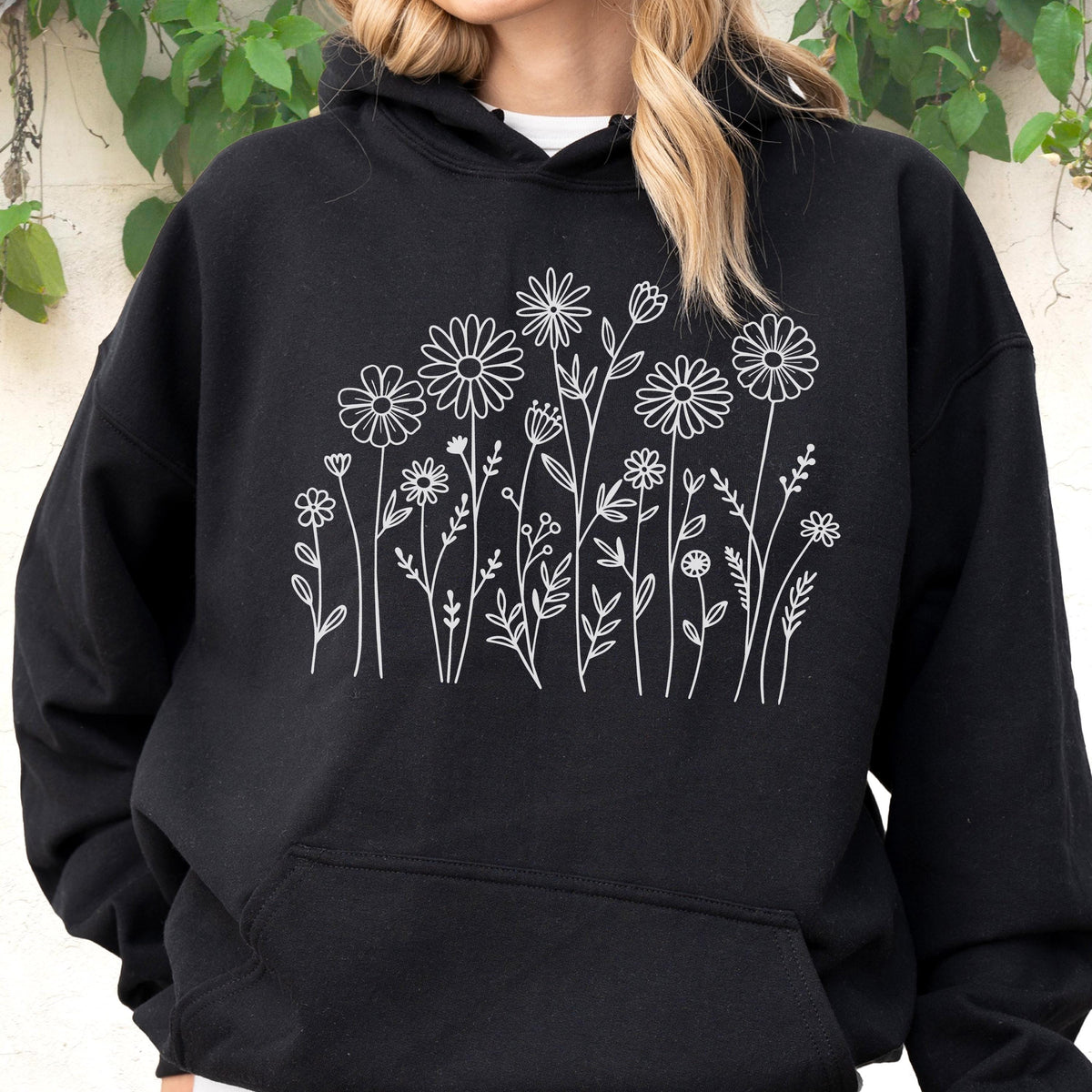 May include: A black hooded sweatshirt featuring a white line art graphic of various wildflowers and stems centered on the chest. The hoodie includes a front kangaroo pocket, long sleeves, and a drawstring hood, providing a simple, clean design.