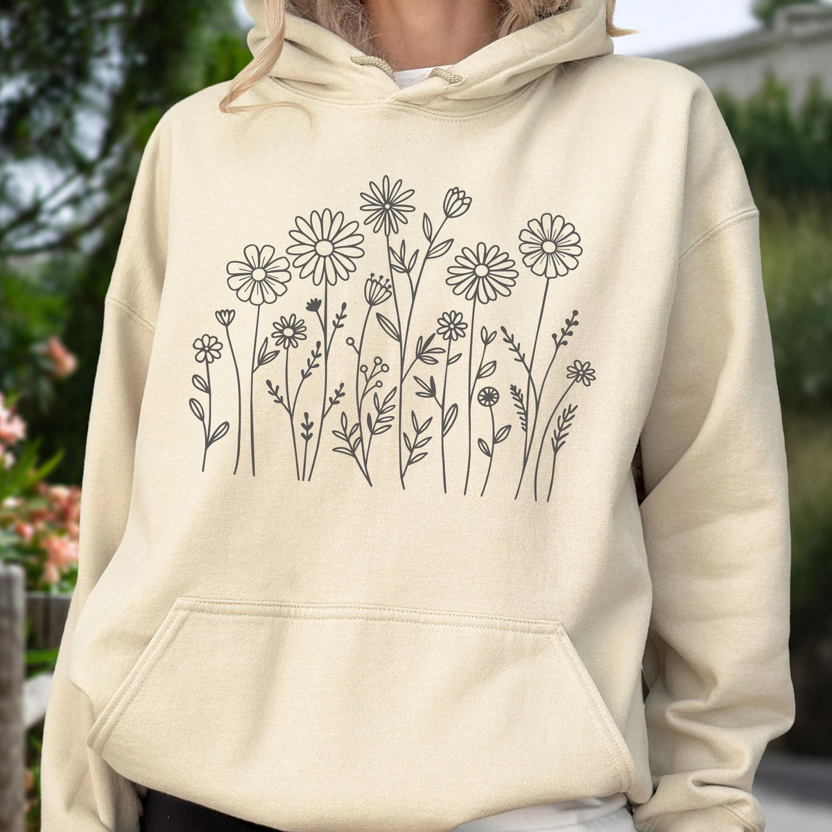 May include: A cream-colored hooded sweatshirt featuring a dark gray line art illustration of various wildflowers and daisies across the chest. The garment includes a front kangaroo pocket, long sleeves, and a drawstring hood with matching cords.