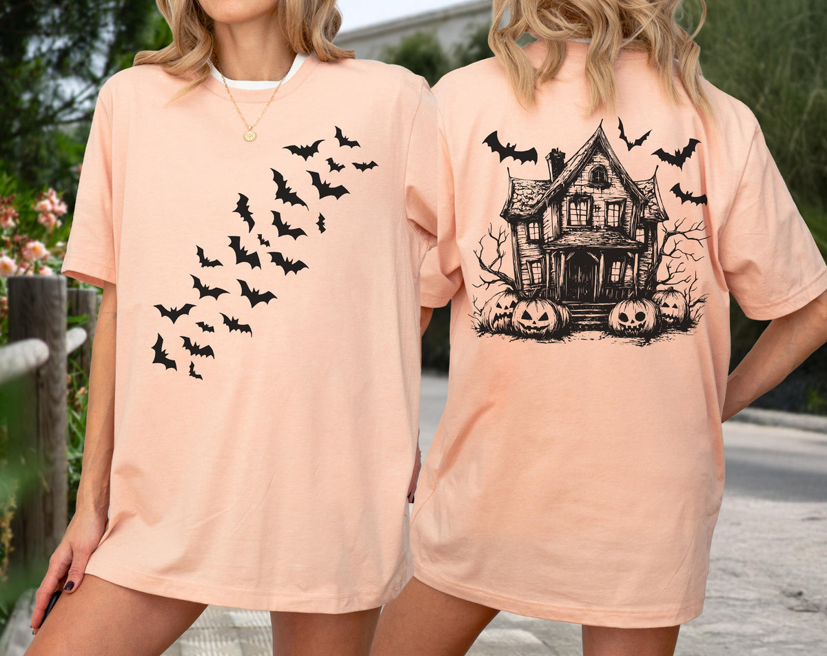 May include: A peach-colored t-shirt featuring a black bat design on the front and a haunted house with pumpkins on the back. The shirt has short sleeves and a crew neck. The design is a Halloween theme.