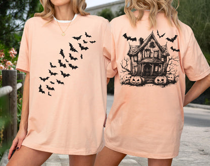 May include: A peach-colored t-shirt featuring a black bat design on the front and a haunted house with pumpkins on the back. The shirt has short sleeves and a crew neck. The design is a Halloween theme.