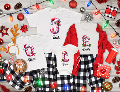 May include: White Christmas-themed family pajamas. The shirts feature candy cane letters with names: Jack, Olivia, Emily, and Liam. The pajamas include a onesie, a long-sleeve shirt, and two short-sleeve shirts. The bottoms are black and white plaid.