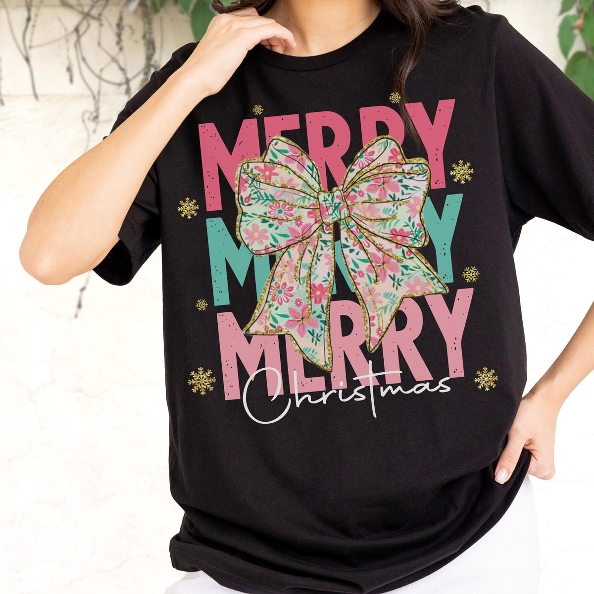 May include: Black t-shirt with the words "MERRY MERRY MERRY Christmas" in pink and teal. A floral bow graphic is centered on the shirt. Gold glitter snowflakes are scattered around the text.