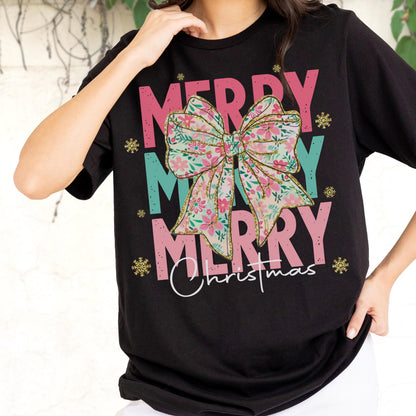 May include: Black t-shirt with the words "MERRY MERRY MERRY Christmas" in pink and teal. A floral bow graphic is centered on the shirt. Gold glitter snowflakes are scattered around the text.